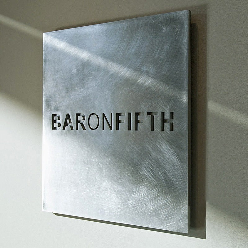 Square stainless steel wall plaque with non-directional vibration finish and precision laser-cut logo "BARONFIFTH" casting architectural shadows