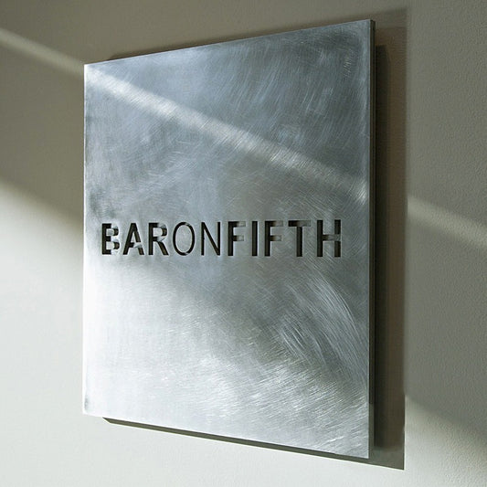 Square stainless steel wall plaque with non-directional vibration finish and precision laser-cut logo "BARONFIFTH" casting architectural shadows