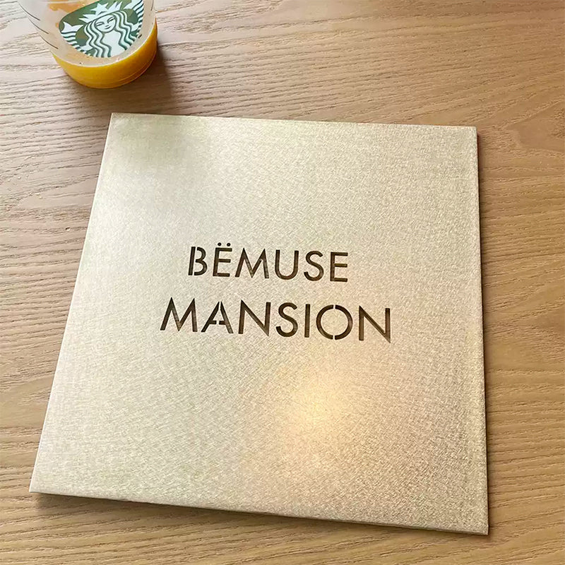 Lifestyle shot of a modern brass sign with laser-cut hollow text ("BEMUSE MANSION"). The cut-out design allows the background surface to show through the letters